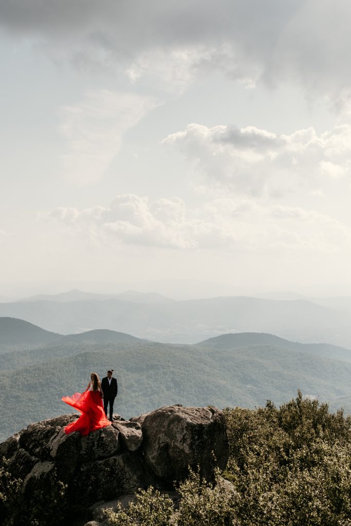 An unconventional bride wearing red, with her groom, stand on top of a mountain ledge to say their vows in an adventure elopement taking place in Virginia's most amazing destination location, the Blue Ridge mountain range. Find out how you can plan an epic elopement incorporating destinatin travel by reading ELOPE