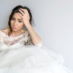 Bride sitting against a wall in her wedding dress, hand on her forehead looking stressed out