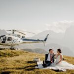 Couple dropped off at their adventure elopement location by a helicopter, having a picnic next to it - with info on When it comes to easy elopement planning, having the right support makes all the difference. Enter the dream team: The Two Pros You Need for Effortless Elopement Planning—your photographer.your Adventure Elopement Photographer and your Adventure Elopement Outfitter.